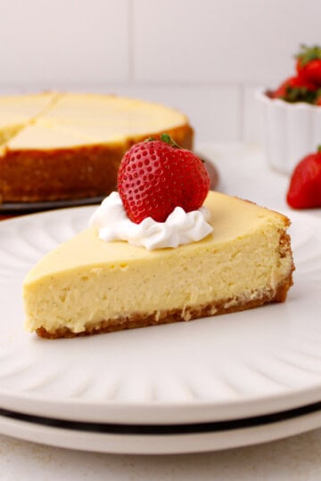 Slice of classic cheesecake on plate with whipped cream and strawberry on top.
